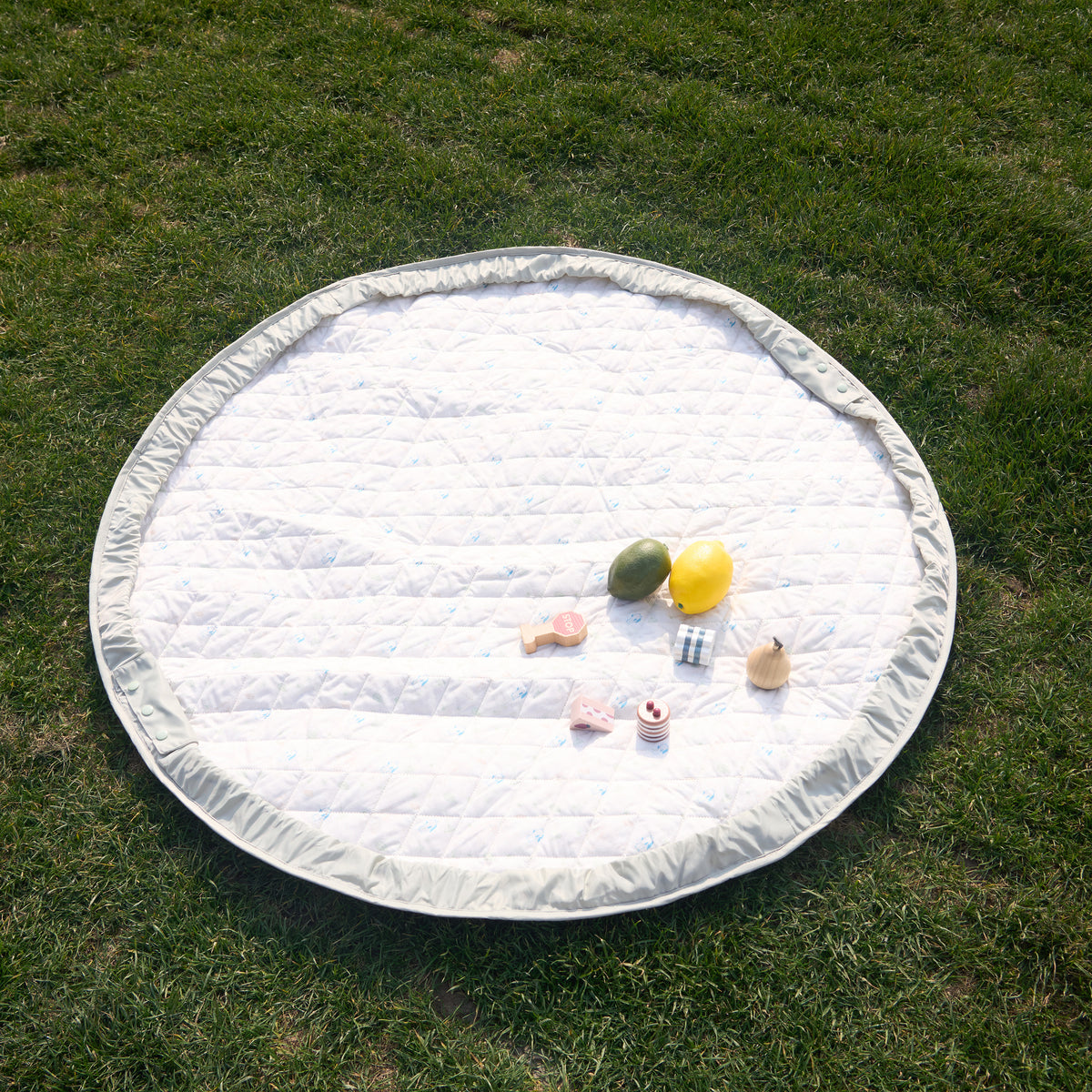 The patterned side of the sage green playmat laid out flat outdoors on green grass, with various baby toys on top.