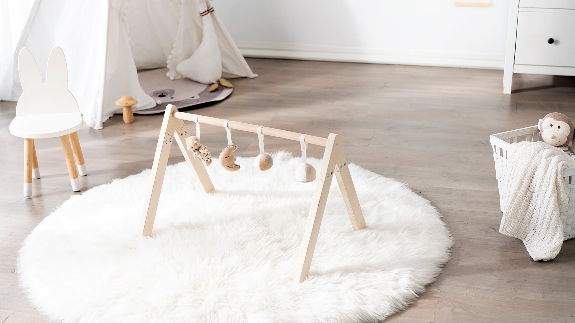 The wooden play gym fully set up in a nursery environment, placed on a white round rug with small dangling toys attached to the crossbar.