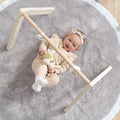 An overhead view of a baby lying comfortably on a round grey playmat, interacting with the wooden activity gym frame overhead.