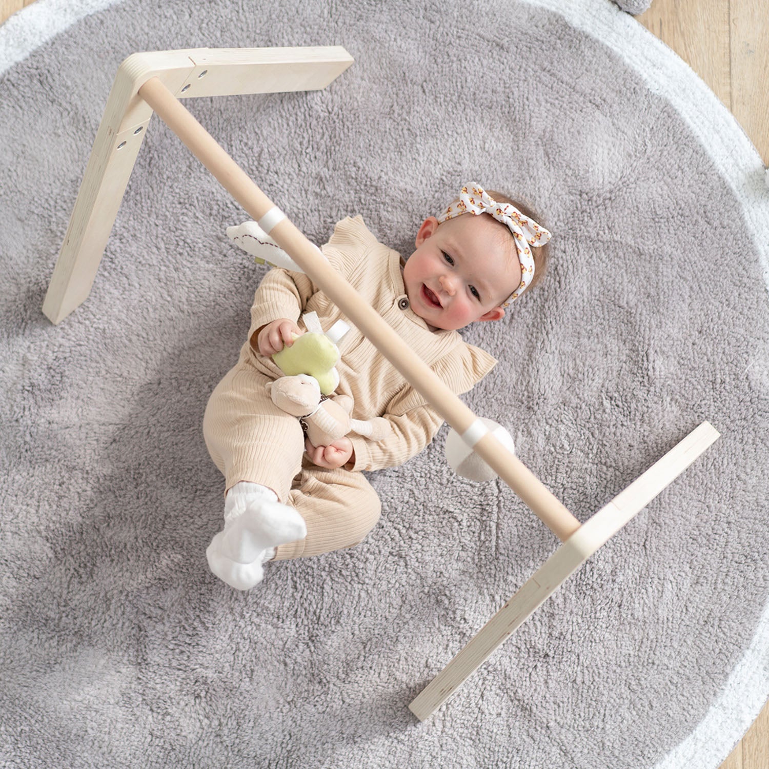 An overhead view of a baby lying comfortably on a round grey playmat, interacting with the wooden activity gym frame overhead.